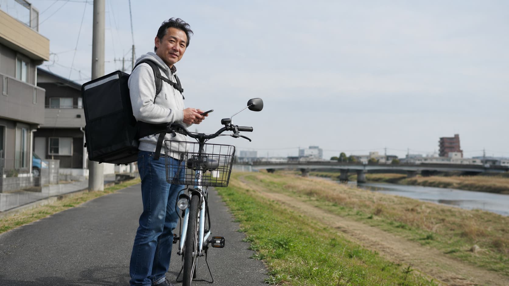 フードデリバリーの配達バッグを背負い自転車のそばに立つ50代の男性