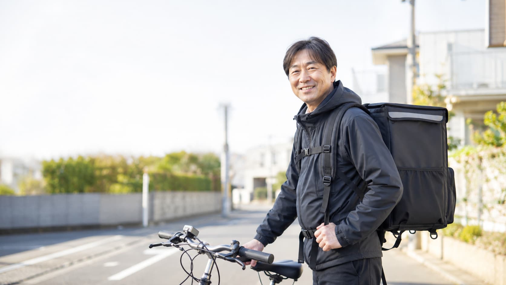 配達用バックパックを背負い、自転車のそばで笑顔を見せる50代の男性